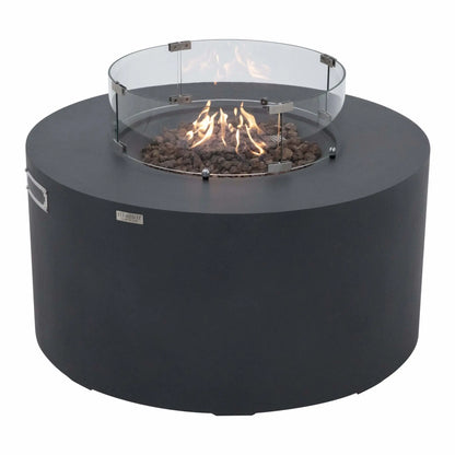 Three-quarter angle view of the Elementi Art of Living Pacaya Dark Grey round GFRC concrete fire pit table operating with visible flames inside the recessed fire bowl. The tempered glass wind guard surrounds the lava rock burner area, highlighting the hidden propane functionality and contemporary outdoor fire feature design.