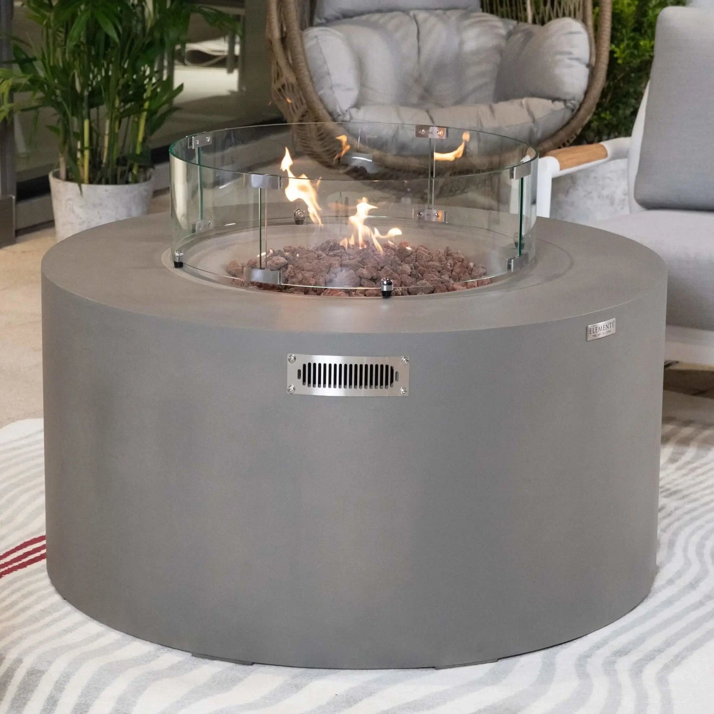 Lifestyle image of the Elementi Art of Living Pacaya Light Grey fire pit table with active flames rising through lava rock media inside a circular tempered glass wind guard. The round GFRC concrete design is showcased in a premium outdoor seating area with modern furniture and greenery, emphasizing warmth, comfort, and upscale outdoor entertaining functionality.