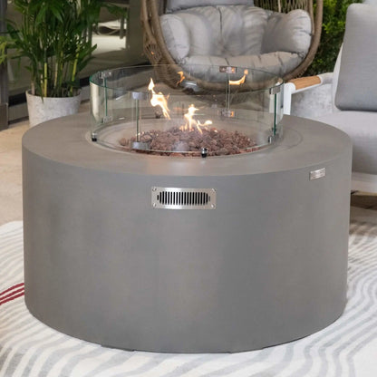 Lifestyle image of the Elementi Art of Living Pacaya Light Grey fire pit table with active flames rising through lava rock media inside a circular tempered glass wind guard. The round GFRC concrete design is showcased in a premium outdoor seating area with modern furniture and greenery, emphasizing warmth, comfort, and upscale outdoor entertaining functionality.