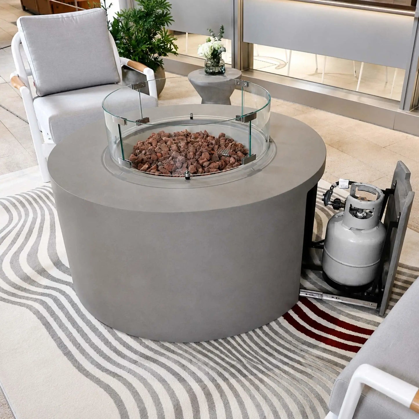 Elementi Art of Living Pacaya Light Grey round outdoor fire pit table shown with a glass wind guard installed and the hidden propane drawer extended to reveal the concealed propane tank storage system. Surrounded by contemporary patio seating, the image demonstrates the convenient hidden propane design, durable GFRC concrete construction, and clean modern outdoor aesthetic.