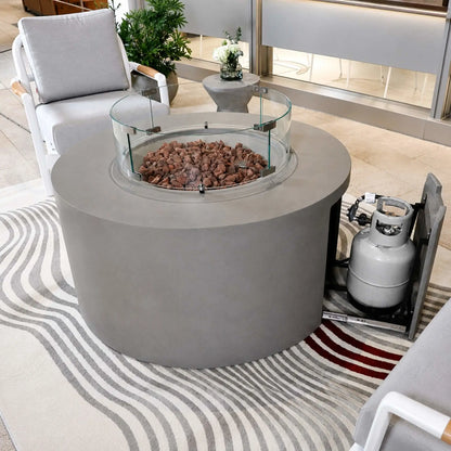 Elementi Art of Living Pacaya Light Grey round outdoor fire pit table shown with a glass wind guard installed and the hidden propane drawer extended to reveal the concealed propane tank storage system. Surrounded by contemporary patio seating, the image demonstrates the convenient hidden propane design, durable GFRC concrete construction, and clean modern outdoor aesthetic.