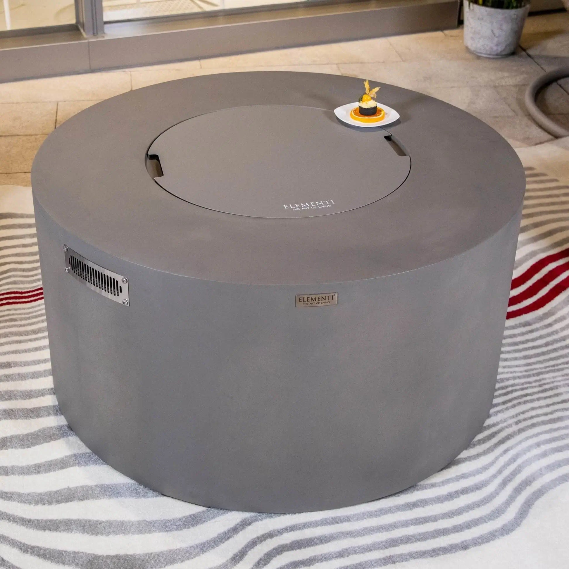 Close-up lifestyle view of the Elementi Art of Living Pacaya Light Grey round fire pit table with the matching burner cover in place. Positioned on a patterned outdoor rug and styled with a decorative dessert plate, the image highlights the smooth GFRC concrete finish, circular tabletop design, and versatility as a functional outdoor coffee table.
