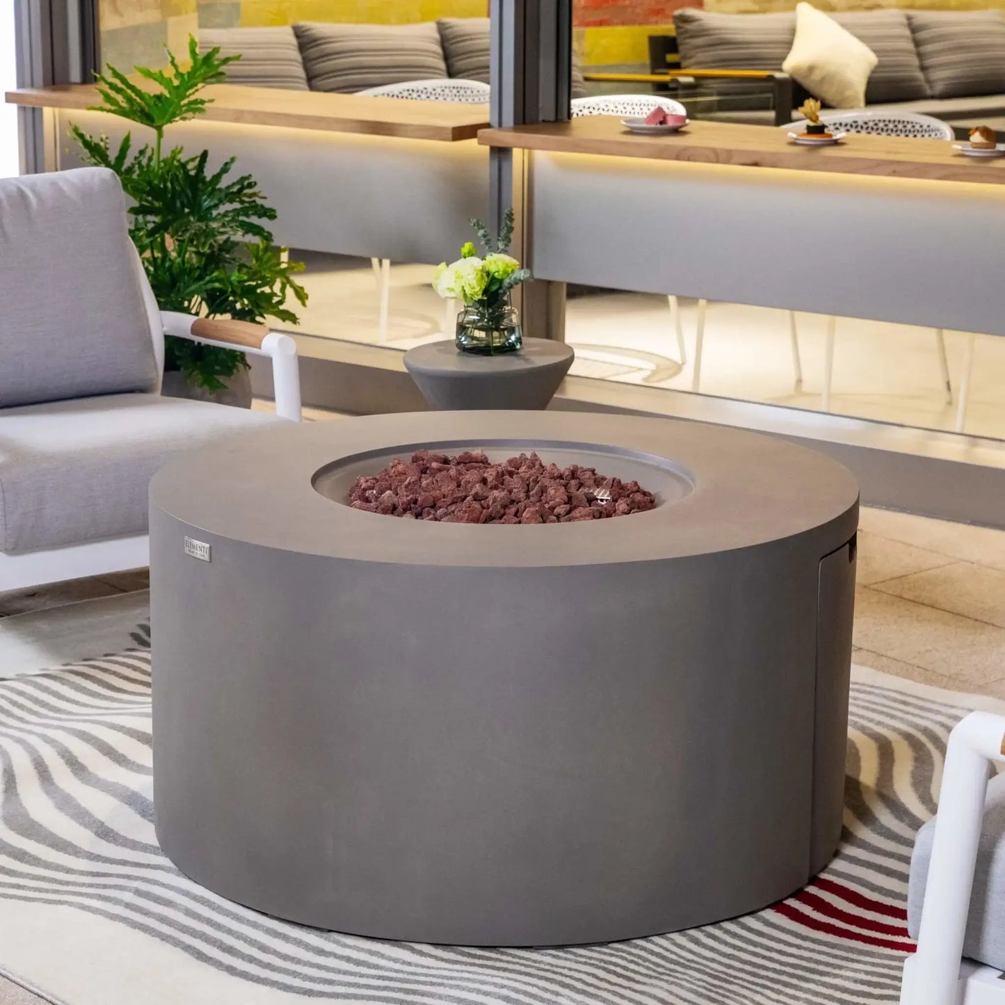 Lifestyle image of the Elementi Art of Living Pacaya Light Grey round GFRC concrete fire pit table with the burner area uncovered and filled with lava rock media. Set within a modern outdoor seating area featuring lounge chairs, plants, and contemporary décor, the image emphasizes the spacious round design and premium outdoor fire feature styling.
