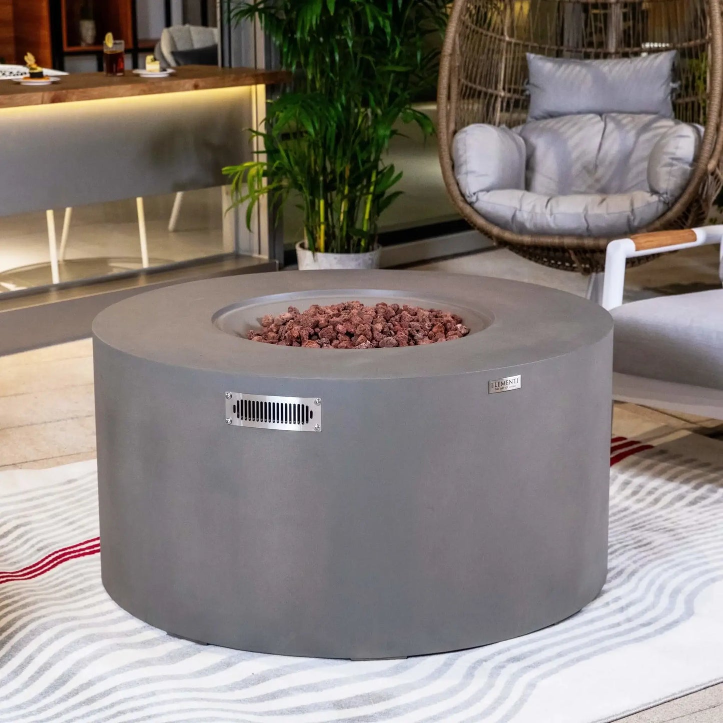 Elementi Art of Living Pacaya Light Grey GFRC concrete fire pit table showcased in a stylish outdoor lounge area with surrounding seating, tropical plants, and decorative furnishings. The recessed lava rock burner area is visible without flames, highlighting the clean circular design, premium concrete construction, and contemporary patio entertainment aesthetic.