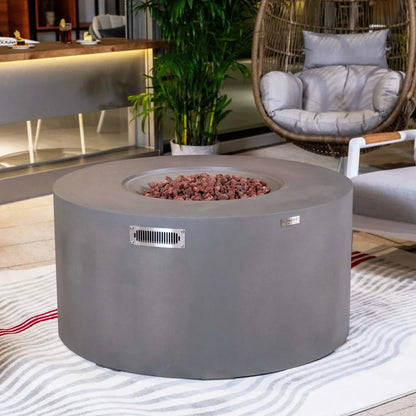 Elementi Art of Living Pacaya Light Grey GFRC concrete fire pit table showcased in a stylish outdoor lounge area with surrounding seating, tropical plants, and decorative furnishings. The recessed lava rock burner area is visible without flames, highlighting the clean circular design, premium concrete construction, and contemporary patio entertainment aesthetic.