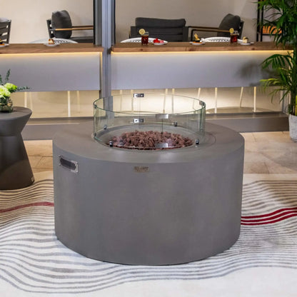 Elementi Art of Living Pacaya Light Grey round outdoor fire pit table fitted with a circular tempered glass wind guard surrounding the lava rock burner area. Positioned within an upscale patio lounge setting, the image showcases the premium GFRC concrete construction, added wind protection, and elegant modern design suitable for outdoor entertaining spaces.