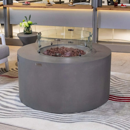 Elementi Art of Living Pacaya Light Grey round GFRC concrete fire pit table displayed on a modern outdoor patio with a circular tempered glass wind guard surrounding the lava rock burner area. Positioned on a patterned outdoor rug and surrounded by contemporary seating and decorative accents, this image highlights the premium concrete construction, wind protection accessory, and sophisticated outdoor entertainment design.