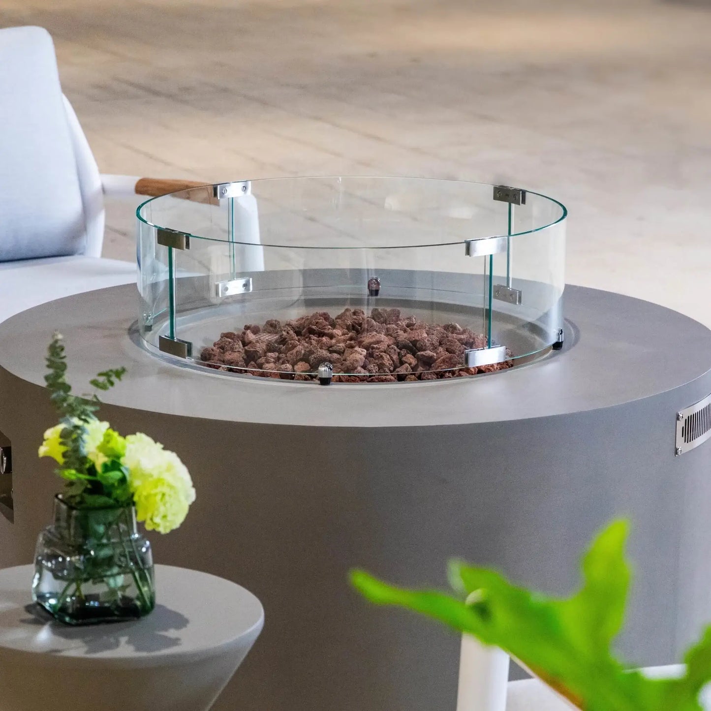 Close-up lifestyle view of the Elementi Art of Living Pacaya Light Grey round fire pit table featuring a clear tempered glass wind guard and natural lava rock media. The image showcases the smooth GFRC concrete finish, circular burner opening, upscale patio furniture, decorative floral arrangement, and refined outdoor living atmosphere ideal for modern gathering spaces.