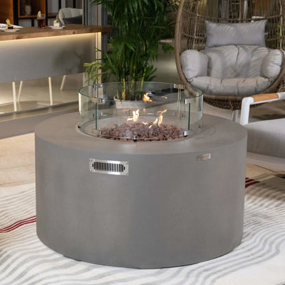 Elementi Art of Living Pacaya Light Grey round GFRC concrete fire pit table operating with visible flames inside a tempered glass wind guard. Set within a stylish outdoor lounge featuring a hanging wicker chair, cushioned seating, and tropical plants, the image highlights the fire feature’s elegant flame presentation, safety glass enclosure, and contemporary patio appeal.