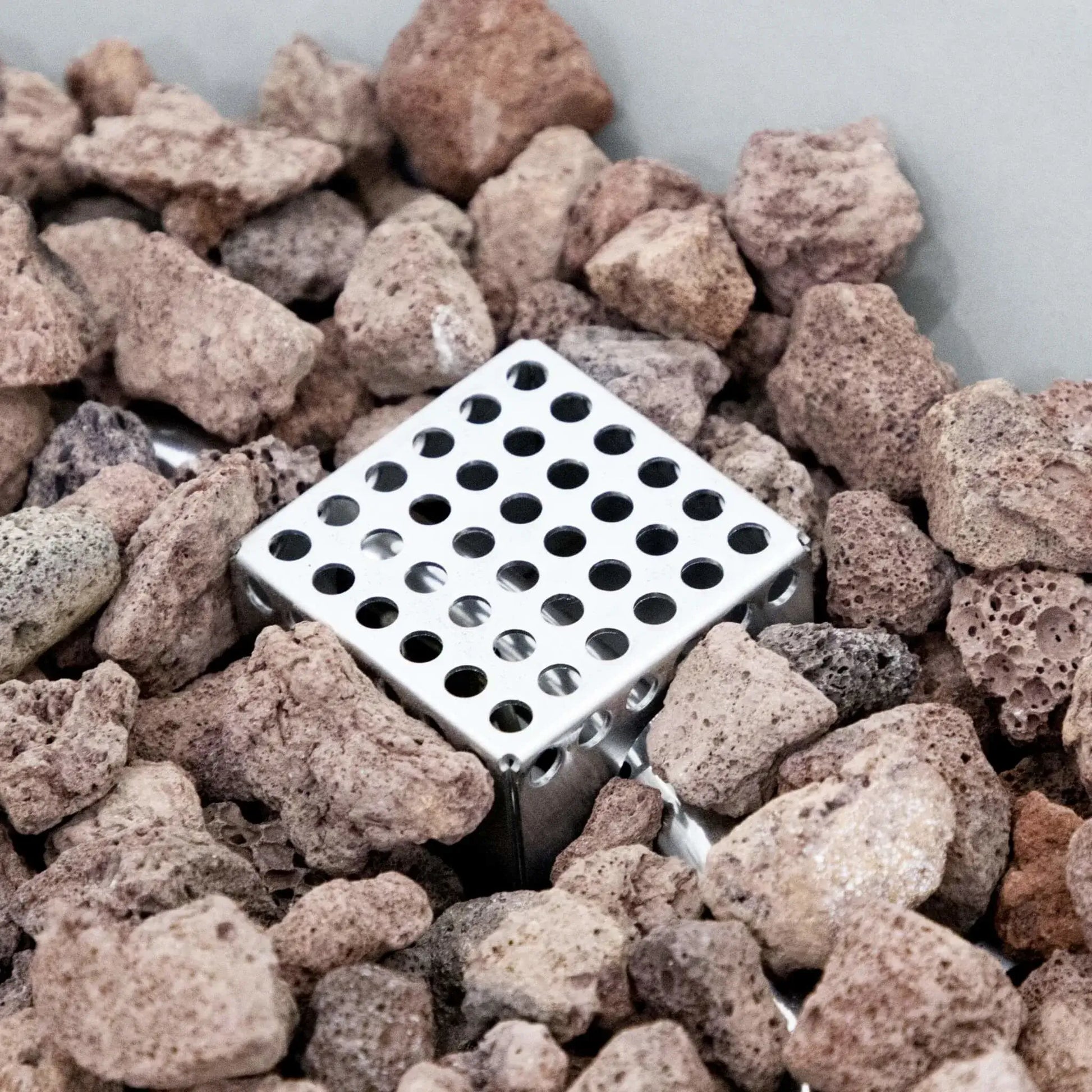 Close-up view of the stainless steel ignition guard and burner component positioned within the lava rock media of the Elementi Art of Living Pacaya Light Grey round GFRC concrete fire pit table. The image highlights the perforated metal ignition housing surrounded by natural volcanic stones, showcasing the concealed burner system and premium construction used in this hidden propane outdoor fire table.