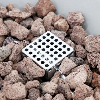 Close-up view of the stainless steel ignition guard and burner component positioned within the lava rock media of the Elementi Art of Living Pacaya Light Grey round GFRC concrete fire pit table. The image highlights the perforated metal ignition housing surrounded by natural volcanic stones, showcasing the concealed burner system and premium construction used in this hidden propane outdoor fire table.