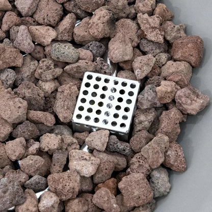 Top-down close-up of the lava rock fire media and stainless steel ignition guard inside the Elementi Art of Living Pacaya Light Grey round fire pit table. The image showcases the natural texture of the volcanic rocks, integrated burner components, and recessed fire bowl design that contribute to the realistic flame presentation and contemporary outdoor aesthetic.