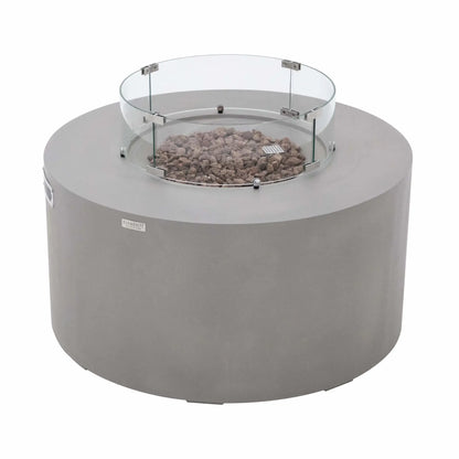 Angled view of the Elementi Art of Living Pacaya round GFRC concrete fire pit table featuring a clear glass wind guard surrounding the lava rock burner area. The image showcases the smooth light grey concrete finish, circular fire bowl design, stainless steel mounting hardware, and contemporary styling that makes the Pacaya fire table a centerpiece for outdoor entertaining spaces.