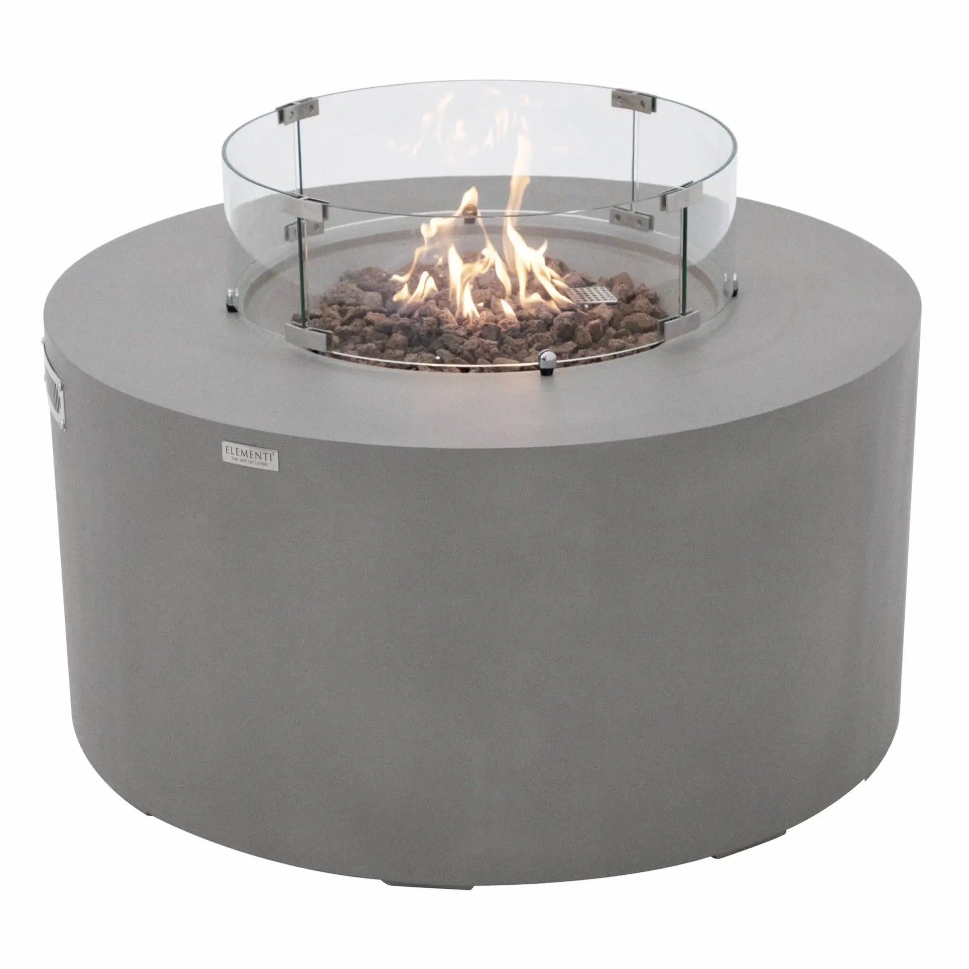 Three-quarter angle view of the Elementi Art of Living Pacaya round fire pit table with the burner lit and protected by a circular glass wind guard. The image showcases dancing flames rising from natural lava rock media, the smooth light grey GFRC concrete body, and the modern low-profile design created for luxury outdoor patios, decks, and backyard gathering areas.