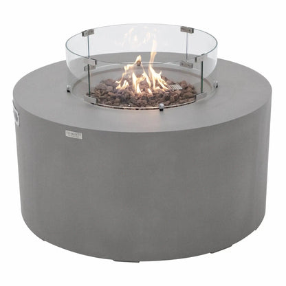 Three-quarter angle view of the Elementi Art of Living Pacaya round fire pit table with the burner lit and protected by a circular glass wind guard. The image showcases dancing flames rising from natural lava rock media, the smooth light grey GFRC concrete body, and the modern low-profile design created for luxury outdoor patios, decks, and backyard gathering areas.