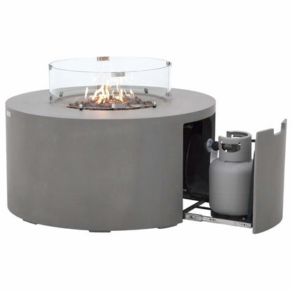 Product image of the Elementi Art of Living Pacaya round fire pit table operating with visible flames while the hidden propane drawer is extended. The slide-out propane tank compartment demonstrates the integrated fuel storage solution, while the tempered glass wind guard, lava rock fire media, and light grey GFRC concrete body showcase the table’s blend of functionality and modern outdoor design.