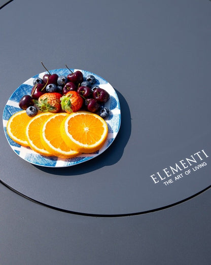 Close-up of the Elementi Pacaya Round Hidden Propane Tank Fire Pit Table in dark grey GFRC concrete, featuring its smooth burner cover elegantly repurposed as a serving surface. A vibrant plate of fresh oranges, cherries, blueberries, and strawberries rests atop, highlighting the table's versatility as both a fire feature and a functional outdoor centerpiece. Perfect for stylish patio gatherings.