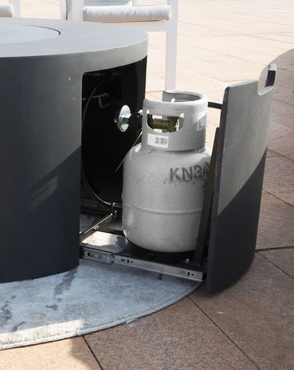 Elementi Pacaya Round Hidden Propane Tank Fire Pit Table in dark grey GFRC concrete, showcasing its innovative pull-out propane tank storage drawer. This sleek outdoor fire feature combines style and function, seamlessly integrating a hidden compartment for easy access and a clean aesthetic. Designed for modern patios, this fire pit table enhances any outdoor space with its minimalist design and effortless usability.