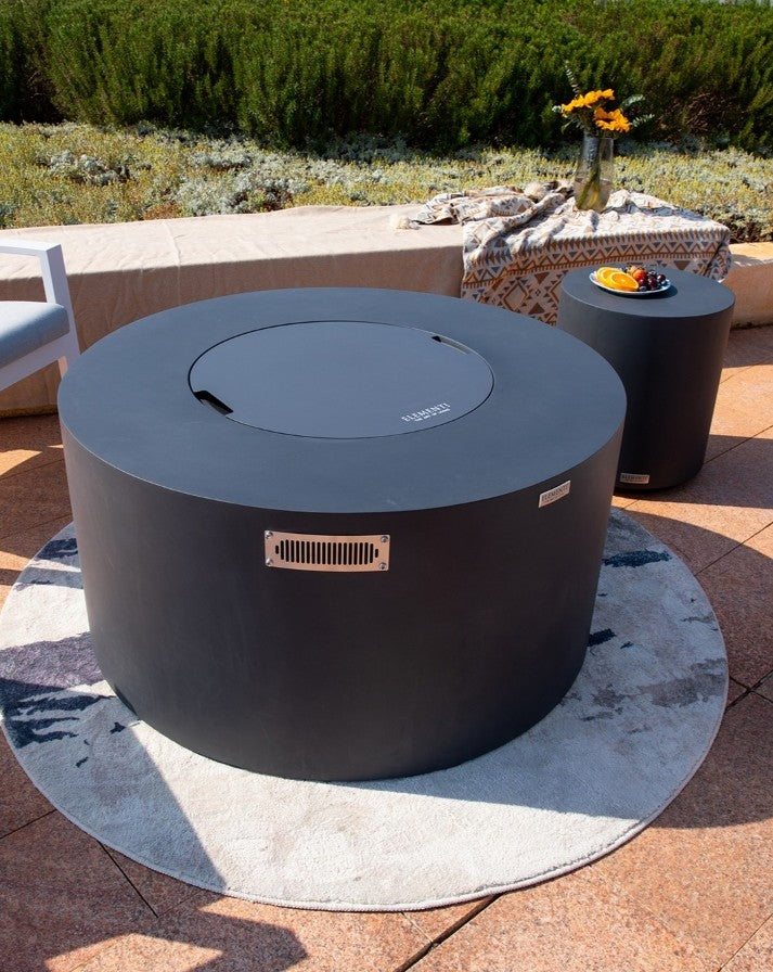 "Elementi Pacaya Round Hidden Propane Tank Fire Pit Table in dark grey GFRC concrete, elegantly positioned in an outdoor setting. The matching side table complements the modern fire feature, creating a sophisticated patio ambiance. A seamless blend of style and function, perfect for luxurious outdoor lounging.
