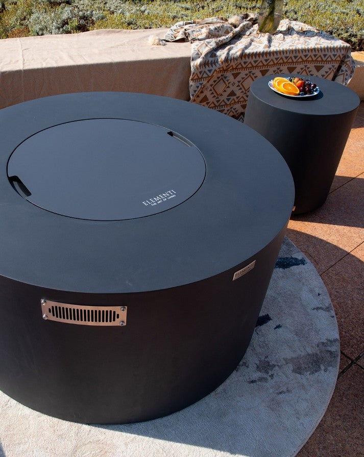 Elementi Pacaya Round Hidden Propane Tank Fire Pit Table in dark grey GFRC concrete with a sleek burner cover, showcased in a stylish outdoor setting. The complementary side table enhances the modern aesthetic, while natural light highlights the smooth concrete finish. A perfect blend of elegance and durability for luxury patio design.