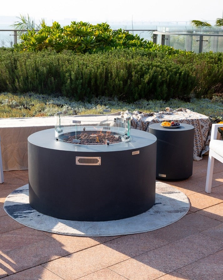 Modern outdoor ambiance with the Elementi Pacaya Round Fire Pit Table in dark grey GFRC concrete, featuring a hidden propane tank and a mesmerizing fire atop lava rocks. The sleek glass wind guard enhances the flame’s elegance, creating a cozy and luxurious patio experience. Paired with a matching side table and stylish seating, this fire feature is perfect for rooftop gatherings or serene backyard retreats.