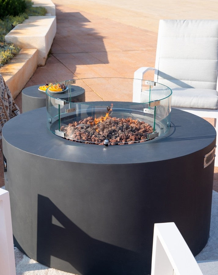 Close-up of the Elementi Pacaya Round Fire Pit Table in dark grey GFRC concrete, featuring a hidden propane tank, vibrant flames dancing over lava rocks, and a sleek glass wind guard. Positioned in a stylish outdoor lounge setting with modern seating and a matching side table, this luxury fire feature creates a warm and inviting atmosphere for elegant patio gatherings.