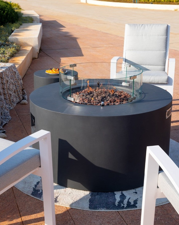 Sophisticated outdoor setting featuring the Elementi Pacaya Round Fire Pit Table in dark grey GFRC concrete. The hidden propane tank design and glass wind guard frame the warm flames flickering over lava rocks, creating an inviting ambiance. Surrounded by modern cushioned seating and a coordinating side table with refreshments, this luxury fire pit is the perfect focal point for stylish patio gatherings.