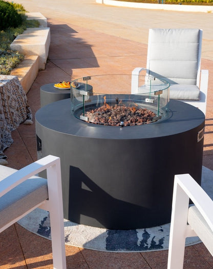 Sophisticated outdoor setting featuring the Elementi Pacaya Round Fire Pit Table in dark grey GFRC concrete. The hidden propane tank design and glass wind guard frame the warm flames flickering over lava rocks, creating an inviting ambiance. Surrounded by modern cushioned seating and a coordinating side table with refreshments, this luxury fire pit is the perfect focal point for stylish patio gatherings.