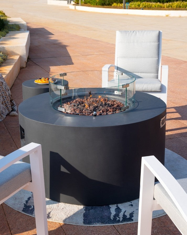 Elegant outdoor lounge featuring the Elementi Pacaya Round Fire Pit Table in dark grey GFRC concrete. With its hidden propane tank, flickering flames over natural lava rocks, and a sleek glass wind guard, this fire feature creates a warm and modern ambiance. Surrounded by contemporary cushioned chairs and a matching side table with refreshments, this setup is perfect for stylish patio gatherings and relaxed evenings under the open sky.
