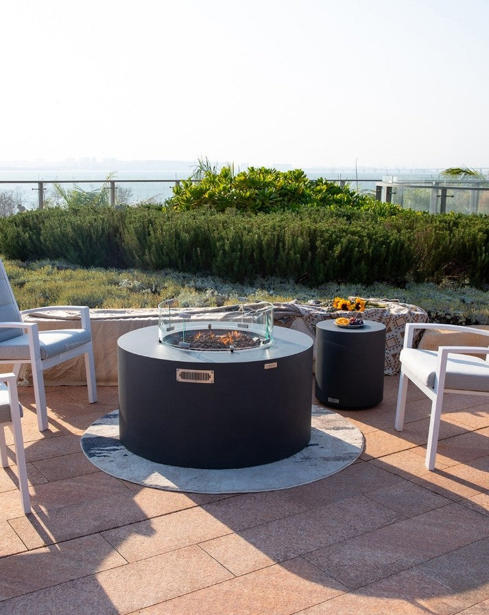 Breathtaking rooftop patio featuring the Elementi Pacaya Round Fire Pit Table in dark grey GFRC concrete. Designed with a hidden propane tank, lava rock media, and a sleek glass wind guard, this fire pit creates a warm and inviting atmosphere. Surrounded by modern cushioned chairs, a matching side table, and scenic greenery, this luxury fire feature is perfect for relaxing evenings and elegant outdoor gatherings.