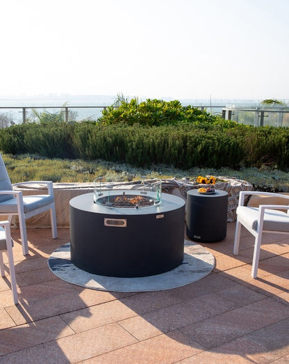 Breathtaking rooftop patio featuring the Elementi Pacaya Round Fire Pit Table in dark grey GFRC concrete. Designed with a hidden propane tank, lava rock media, and a sleek glass wind guard, this fire pit creates a warm and inviting atmosphere. Surrounded by modern cushioned chairs, a matching side table, and scenic greenery, this luxury fire feature is perfect for relaxing evenings and elegant outdoor gatherings.