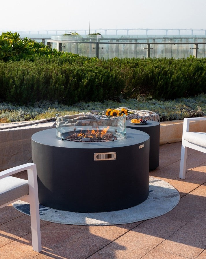 Luxurious outdoor retreat featuring the Elementi Pacaya Round Fire Pit Table in dark grey GFRC concrete. With its hidden propane tank, flickering flames over natural lava rocks, and a contemporary glass wind guard, this fire feature creates a warm and elegant ambiance. Set against a scenic backdrop with modern seating, a matching side table, and decorative accents, this patio setup is perfect for upscale gatherings and cozy evenings under the sky.