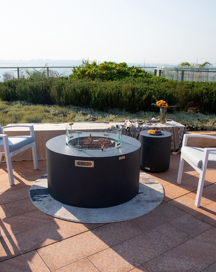 Elementi Pacaya Round Hidden Propane Tank Fire Pit Table in dark grey GFRC concrete, elegantly set on a scenic outdoor patio. Featuring a protective glass wind guard and natural lava rocks, this fire pit creates a cozy and sophisticated ambiance. Its hidden propane tank compartment ensures a sleek, modern aesthetic. Surrounded by stylish seating and complemented by a matching side table adorned with fresh fruit, this luxury fire feature is the perfect statement piece for any upscale outdoor retreat.