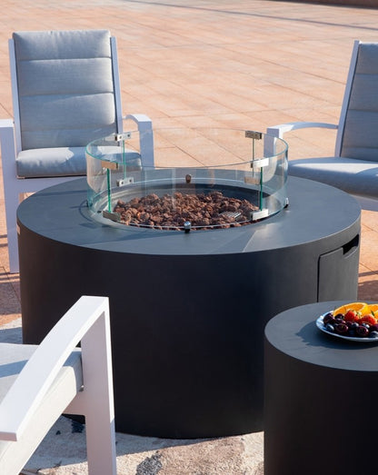 "Elementi Pacaya Round Hidden Propane Tank Fire Pit Table in dark grey GFRC concrete, featuring a glass wind guard and natural lava rock for a refined outdoor ambiance. The seamless design integrates a hidden propane tank compartment for a clean, modern aesthetic. Complemented by a matching side table and surrounded by sleek contemporary chairs, this luxury fire feature elevates any patio or outdoor lounge space with both style and warmth.