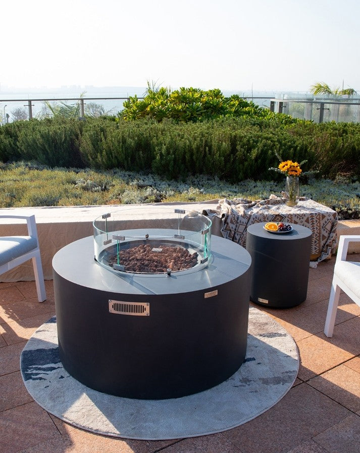 Stylish outdoor setup featuring the Elementi Pacaya Round Fire Pit Table in dark grey GFRC concrete. The sleek, modern design includes a hidden propane tank, a glass wind guard, and lava rock media for a warm, inviting ambiance. Complemented by a matching side table and cozy seating, this elegant fire feature transforms any patio or rooftop into a luxurious retreat with stunning scenic views.