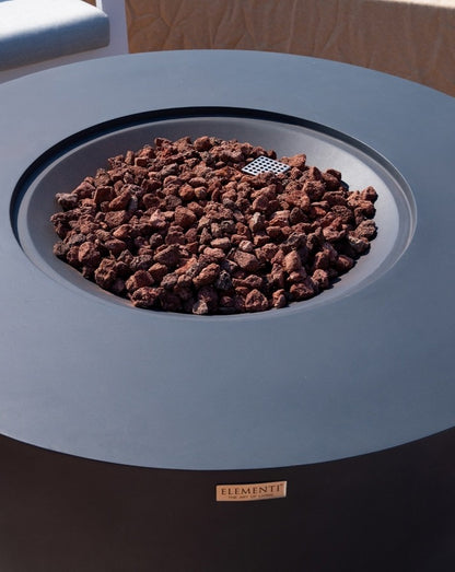 Close-up of the Elementi Pacaya Round Hidden Propane Tank Fire Pit Table in dark grey GFRC concrete, showcasing rich, natural lava rock and a precision-engineered burner. The sleek, modern design highlights the fine craftsmanship and durable materials, creating a stunning focal point for outdoor living spaces. Perfect for adding warmth and ambiance to any contemporary patio or luxury terrace.