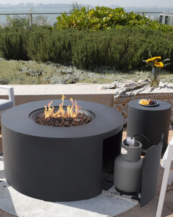 Elementi Pacaya Round Hidden Propane Tank Fire Pit Table in dark grey GFRC concrete, elegantly ignited with vibrant flames over natural lava rock. The seamless hidden propane tank design ensures a clean, modern aesthetic, while the coordinating side table enhances the luxurious outdoor ambiance. Set against a stunning natural backdrop, this fire pit is the ultimate statement piece for sophisticated patio and rooftop lounging.
