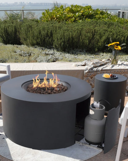 Elementi Pacaya Round Hidden Propane Tank Fire Pit Table in dark grey GFRC concrete, elegantly ignited with vibrant flames over natural lava rock. The seamless hidden propane tank design ensures a clean, modern aesthetic, while the coordinating side table enhances the luxurious outdoor ambiance. Set against a stunning natural backdrop, this fire pit is the ultimate statement piece for sophisticated patio and rooftop lounging.