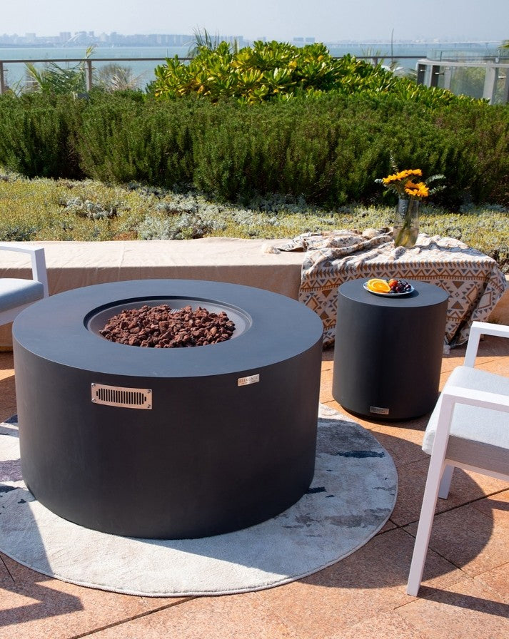 Elementi Pacaya Round Hidden Propane Tank Fire Pit Table in dark grey GFRC concrete, elegantly styled in an outdoor setting with a coordinating side table. Featuring natural lava rock for an authentic fire experience, this modern fire pit blends seamlessly into contemporary patios and luxury terraces. The sleek design and rich textures create a cozy, sophisticated ambiance, perfect for entertaining or relaxing under the open sky.