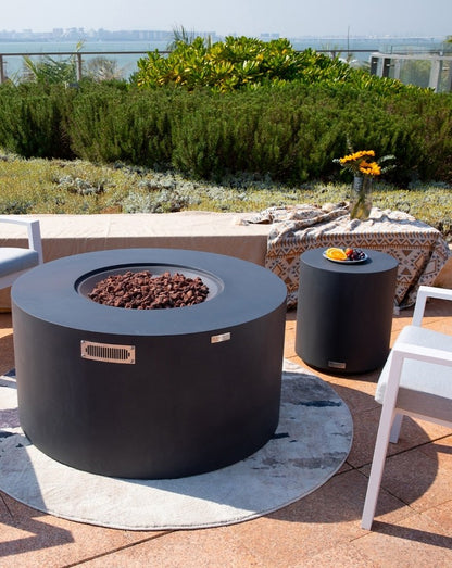 Elementi Pacaya Round Hidden Propane Tank Fire Pit Table in dark grey GFRC concrete, elegantly styled in an outdoor setting with a coordinating side table. Featuring natural lava rock for an authentic fire experience, this modern fire pit blends seamlessly into contemporary patios and luxury terraces. The sleek design and rich textures create a cozy, sophisticated ambiance, perfect for entertaining or relaxing under the open sky.