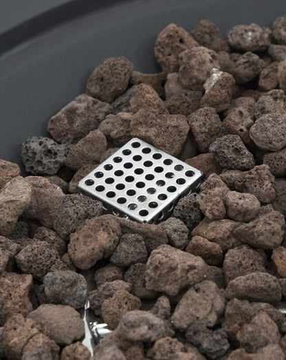 Detailed close-up of the Elementi Pacaya Round Fire Pit Table’s stainless steel gas diffuser, surrounded by natural lava rocks. Designed for optimal heat distribution and an even flame output, the perforated burner component enhances efficiency and performance, creating a warm and stylish outdoor fire feature with a modern touch.