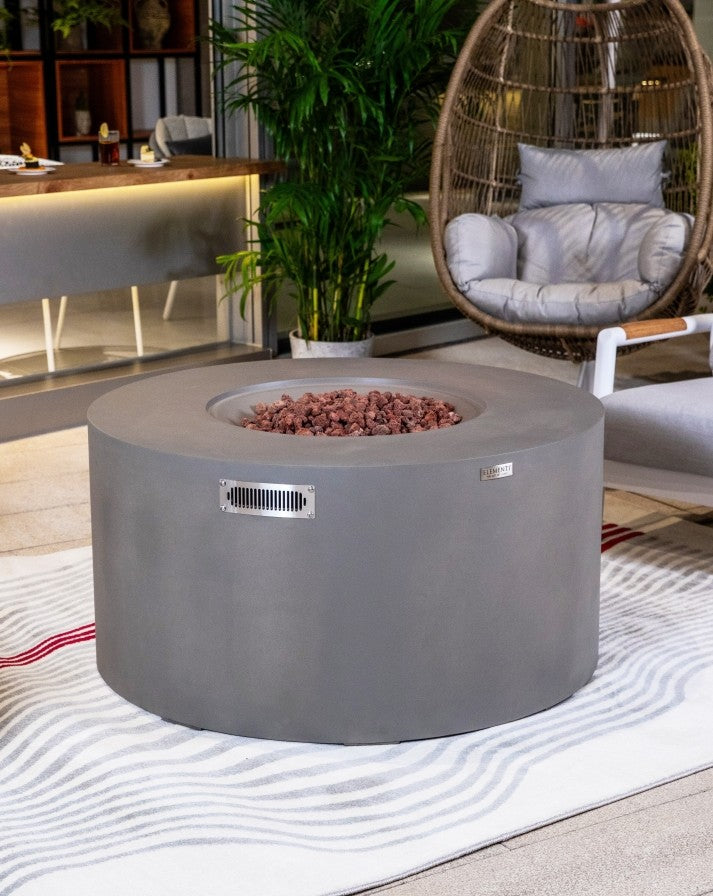 Elementi Pacaya Round Hidden Propane Tank Fire Pit Table in light grey GFRC concrete, elegantly placed in a modern outdoor lounge, featuring natural lava rocks and a sleek ventilation panel for both style and functionality.