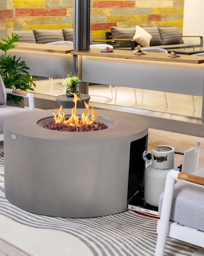 Elementi Pacaya Round Hidden Propane Tank Fire Pit Table in light grey GFRC concrete, featuring a mesmerizing flame over natural lava rocks, with a discreet pull-out propane tank storage drawer for seamless functionality and modern outdoor elegance.