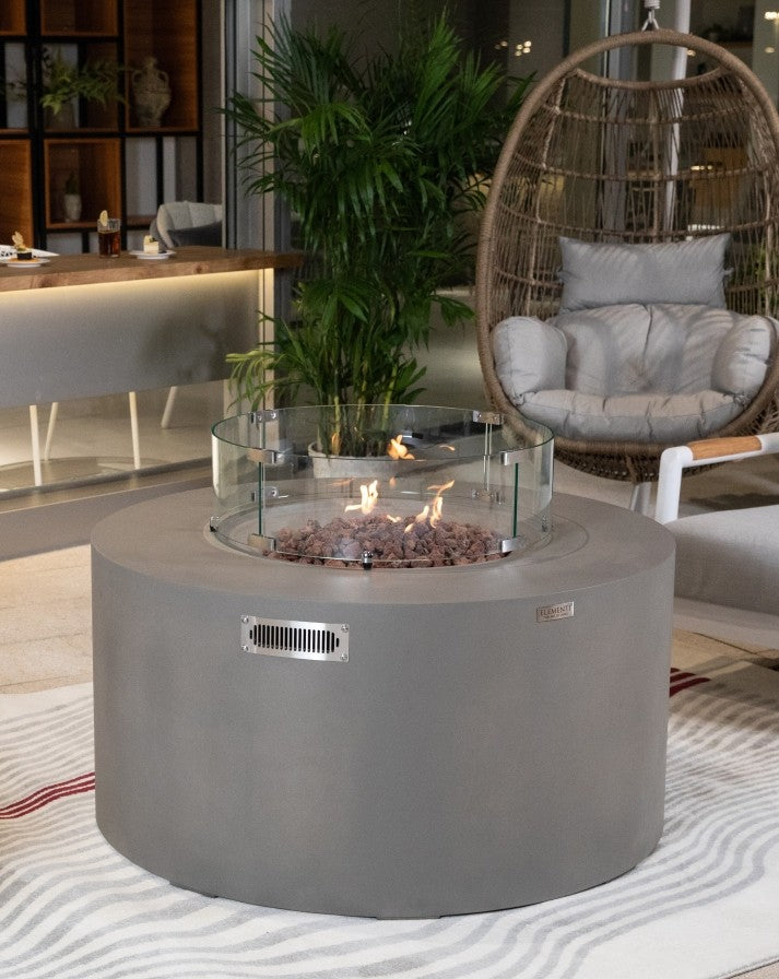 Elementi Pacaya Round Hidden Propane Tank Fire Pit Table in light grey GFRC concrete, featuring a mesmerizing flame over natural lava rocks, elegantly enclosed by a glass wind guard, creating a cozy and stylish outdoor retreat.