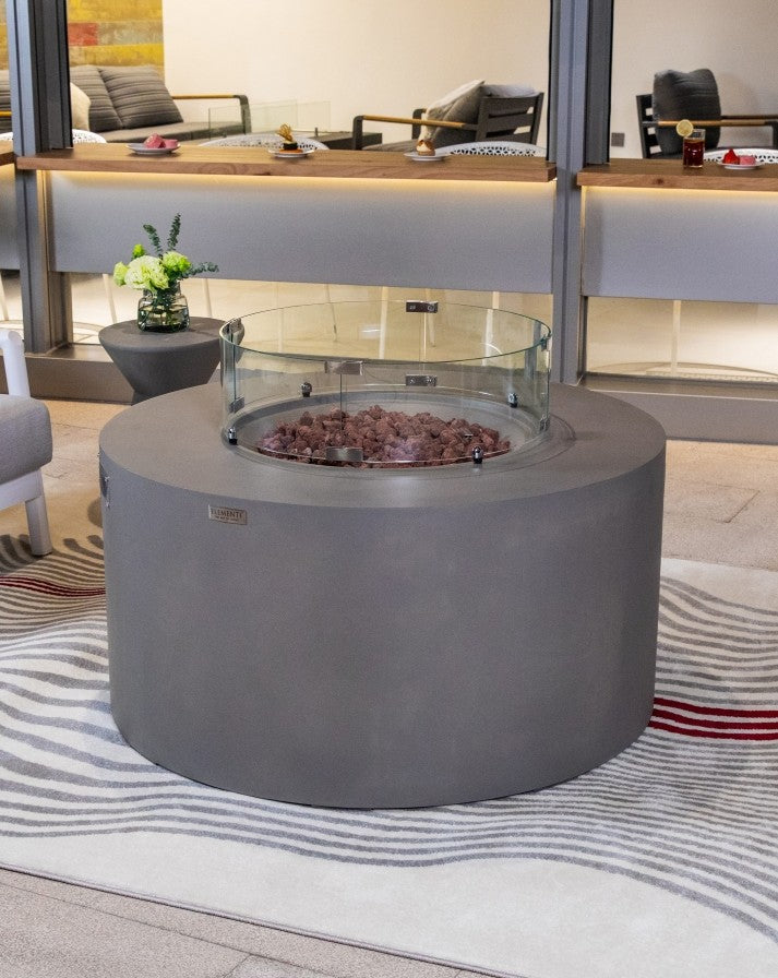 Elementi Pacaya Round Hidden Propane Tank Fire Pit Table in light grey GFRC concrete, featuring a glass wind guard and natural lava rocks, seamlessly blending modern design with cozy outdoor ambiance.