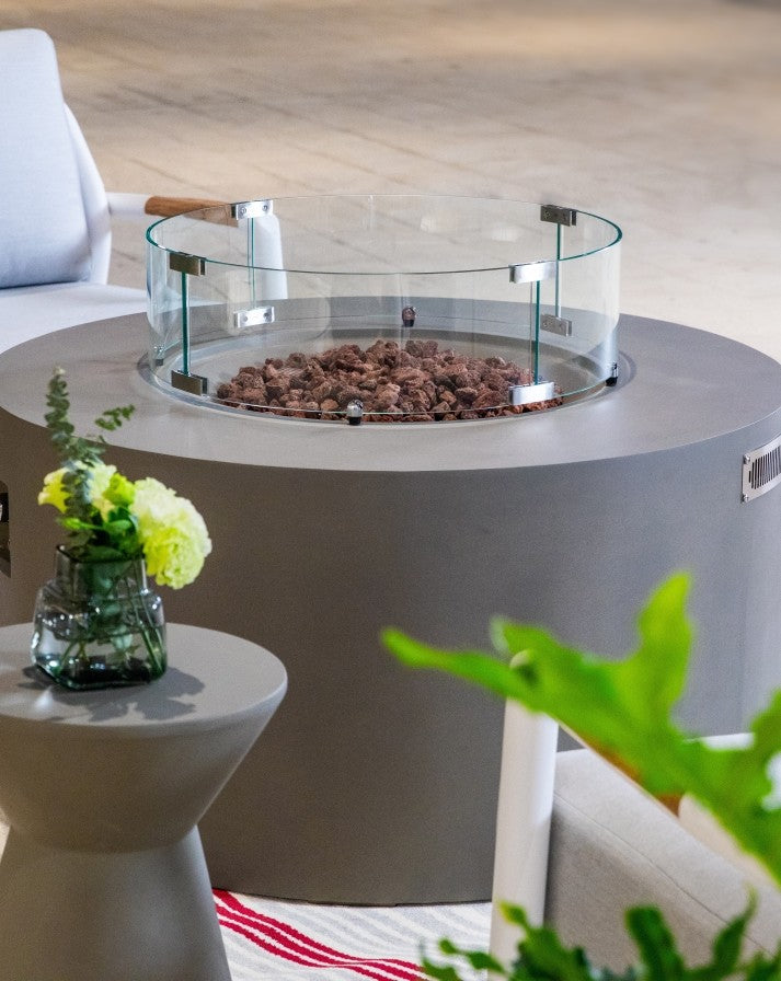 Close-up of the Elementi Pacaya Round Hidden Propane Tank Fire Pit Table in light grey GFRC concrete, featuring a sleek glass wind guard and natural lava rocks, adding a modern and sophisticated touch to any outdoor setting.