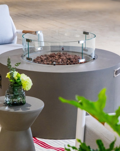 Close-up of the Elementi Pacaya Round Hidden Propane Tank Fire Pit Table in light grey GFRC concrete, featuring a sleek glass wind guard and natural lava rocks, adding a modern and sophisticated touch to any outdoor setting.