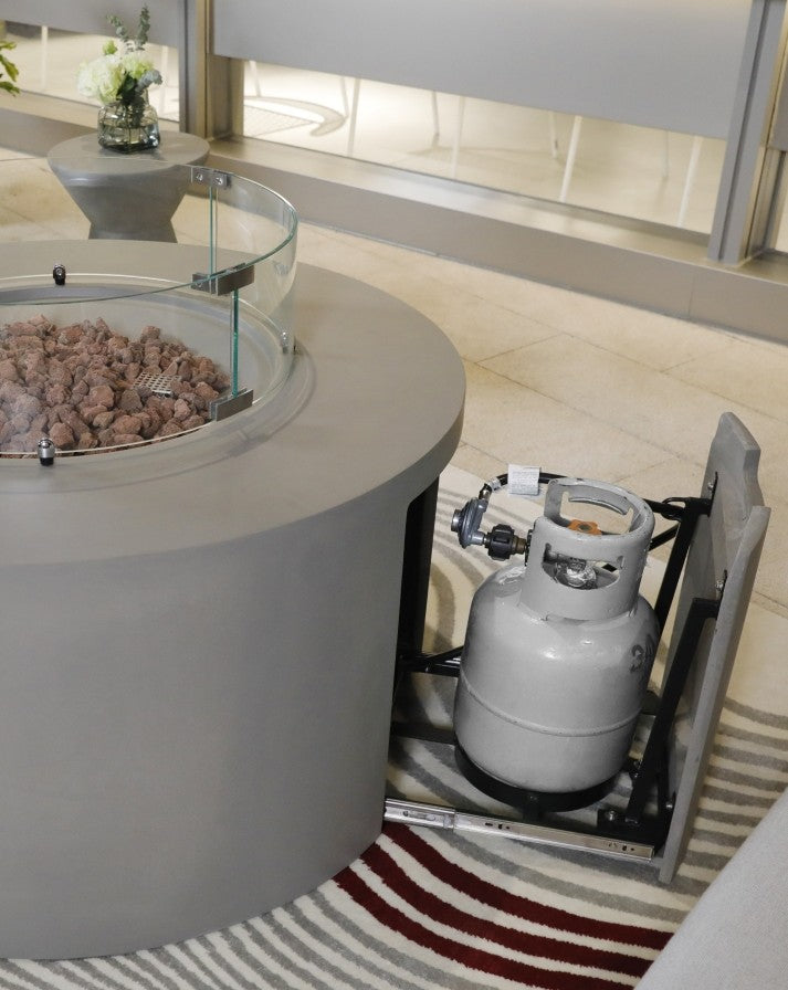 Elementi Pacaya Round Hidden Propane Tank Fire Pit Table in light grey GFRC concrete, featuring a sleek glass wind guard, natural lava rocks, and a discreet pull-out propane tank storage drawer for a seamless and stylish outdoor experience.