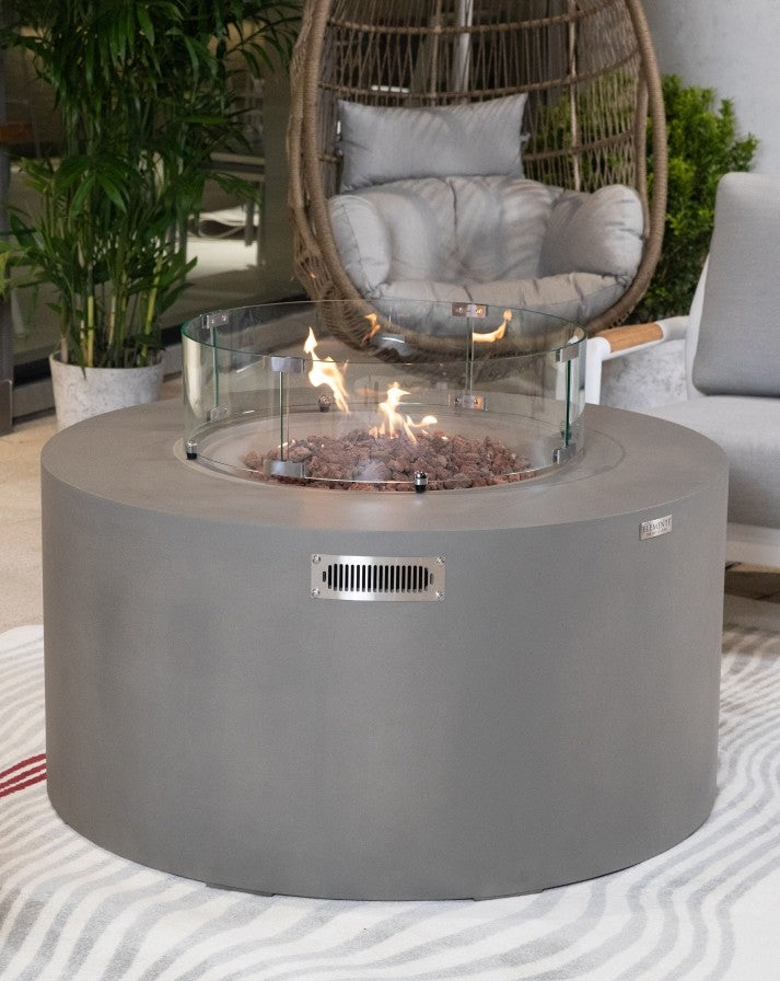 Elementi Pacaya Round Hidden Propane Tank Fire Pit Table in light grey GFRC concrete, featuring a warm, flickering flame over natural lava rocks, protected by a sleek glass wind guard, creating a luxurious and inviting outdoor ambiance.