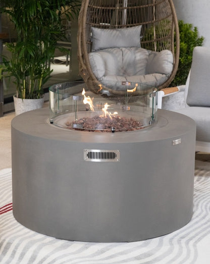 Elementi Pacaya Round Hidden Propane Tank Fire Pit Table in light grey GFRC concrete, featuring a warm, flickering flame over natural lava rocks, protected by a sleek glass wind guard, creating a luxurious and inviting outdoor ambiance.